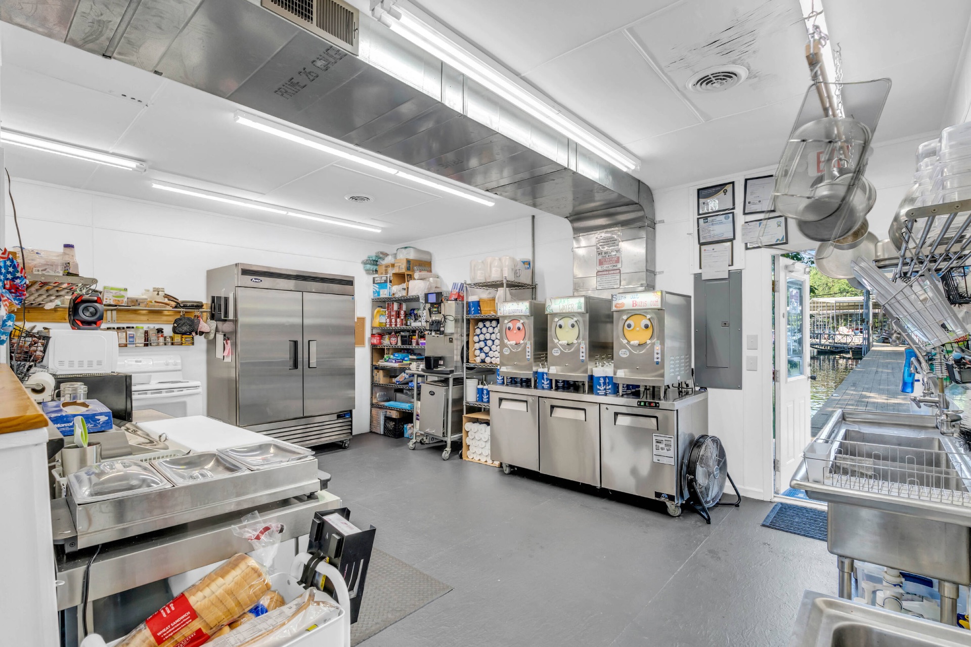 Marina kitchen with three frozen slushie machines, prep counters, refrigeration, and a doorway out to the dock.